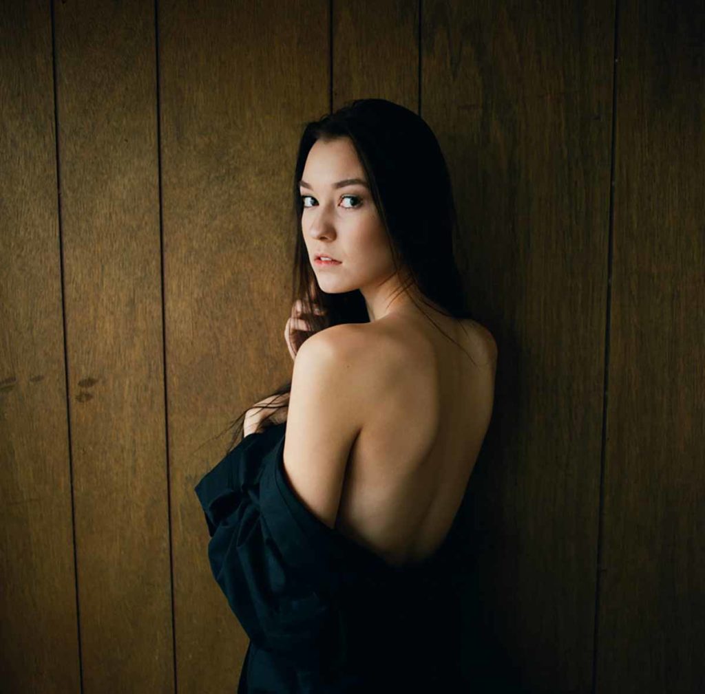 Photo. A young woman in front of a wall, her back to the viewer with her robe top oulled down to expose her back. She looks over her shoulder at the viewer.