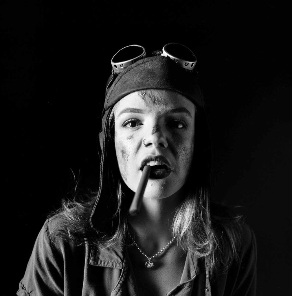 A black and white photo of a young woman in a cap with goggles on top of her head, a cigar in her mouth, and grease on her face, looking defiantly into the camera.