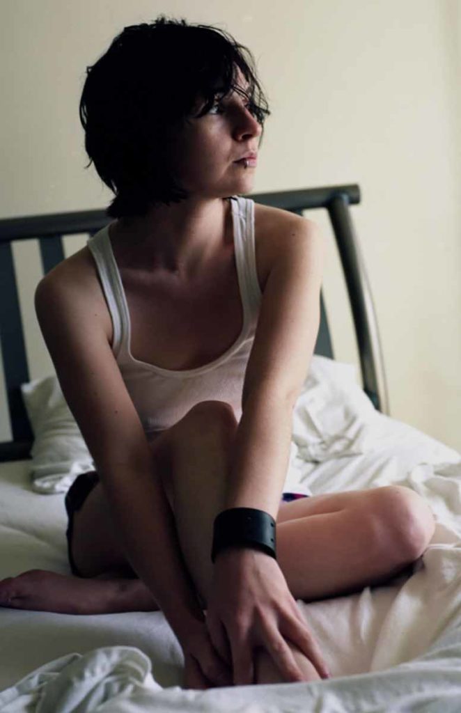 Photo. A young woman sitting on a bed, looking away from the viewer.