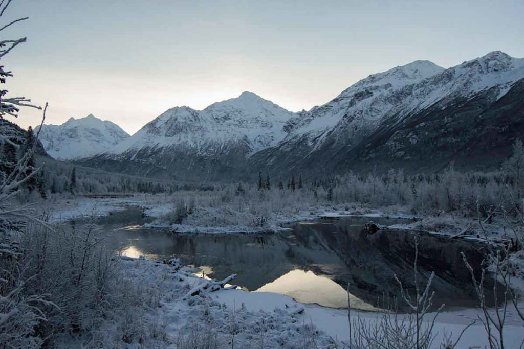 Photo. A winter scene in Alaska of a lake surrounded by frost-covered trees with majestic mountains in the background.