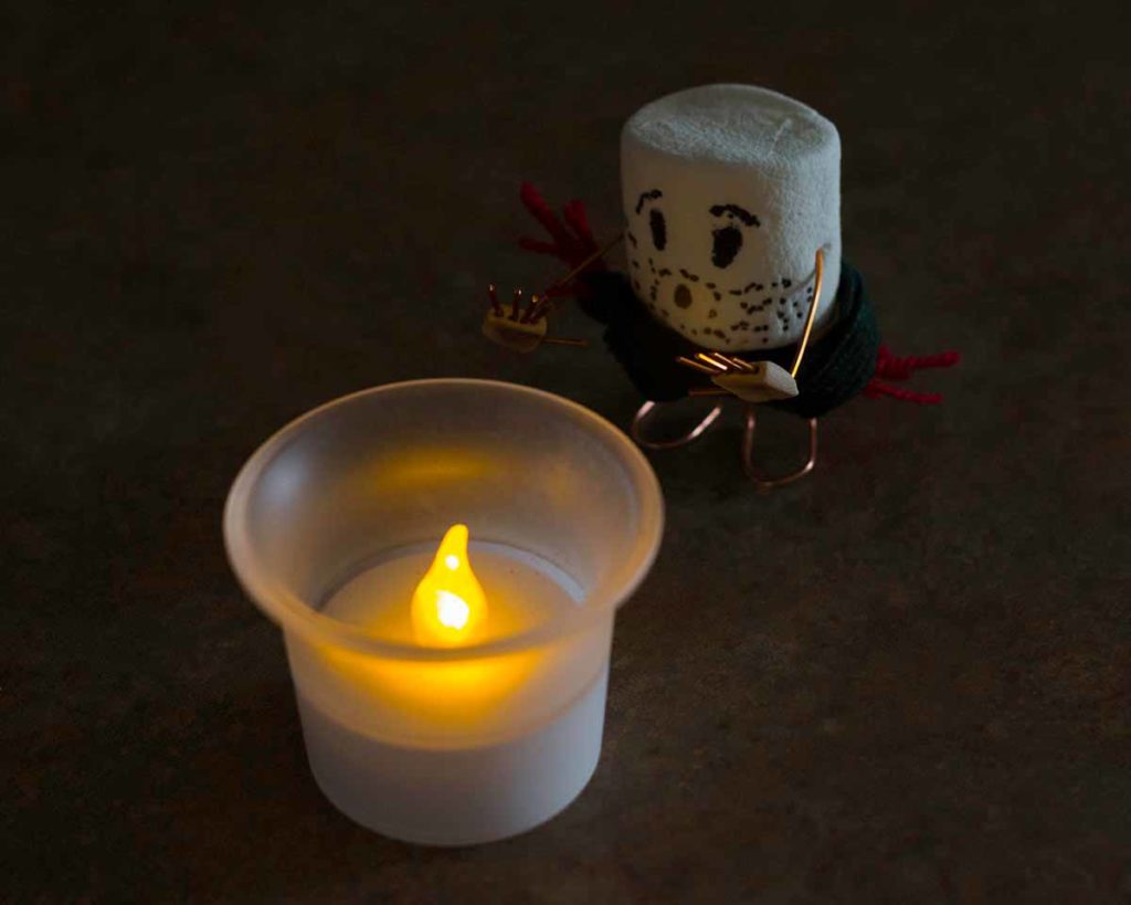 Photo. An image of a marshmallow with a face drawn on it and copper limbs, warming its hands on the flame from a votive candle.