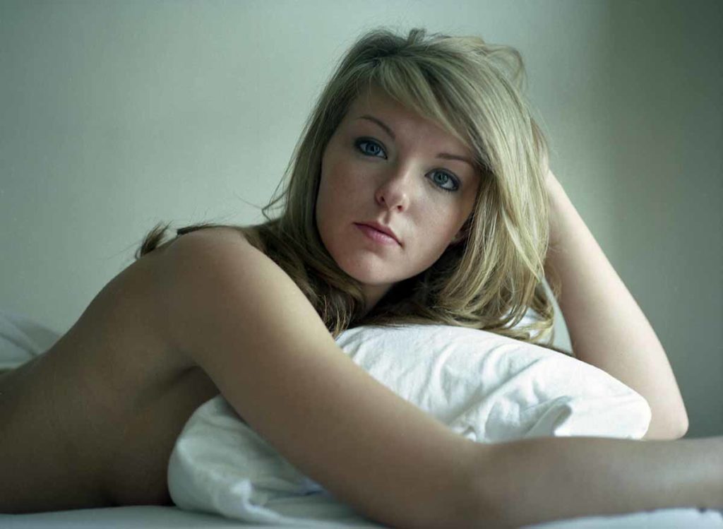 Photo. A young womanlaying face down on a bed, head propped up looking at the viewer.