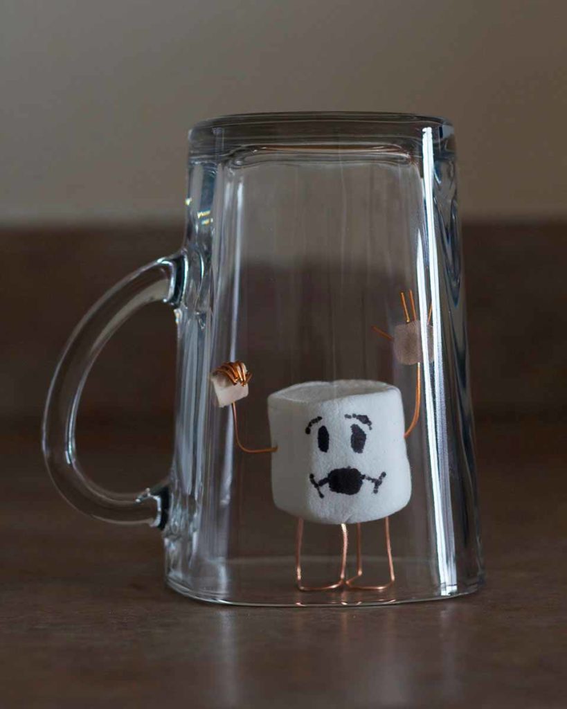 Photo. A playful scene of a marshmallow with a drawon on face and copper wire limbs, trapped under an upside-down glass.