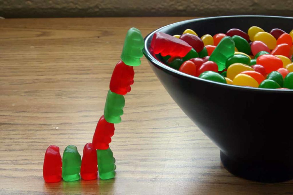 Photo. A playful image of a group of gummy bear candies forming a ladder to get into a candy dish full of jelly beans.